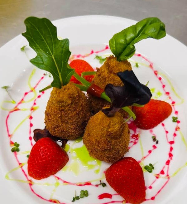 A white plate holds golden-brown fried pakora surrounded by strawberry halves and decorative sauces.
